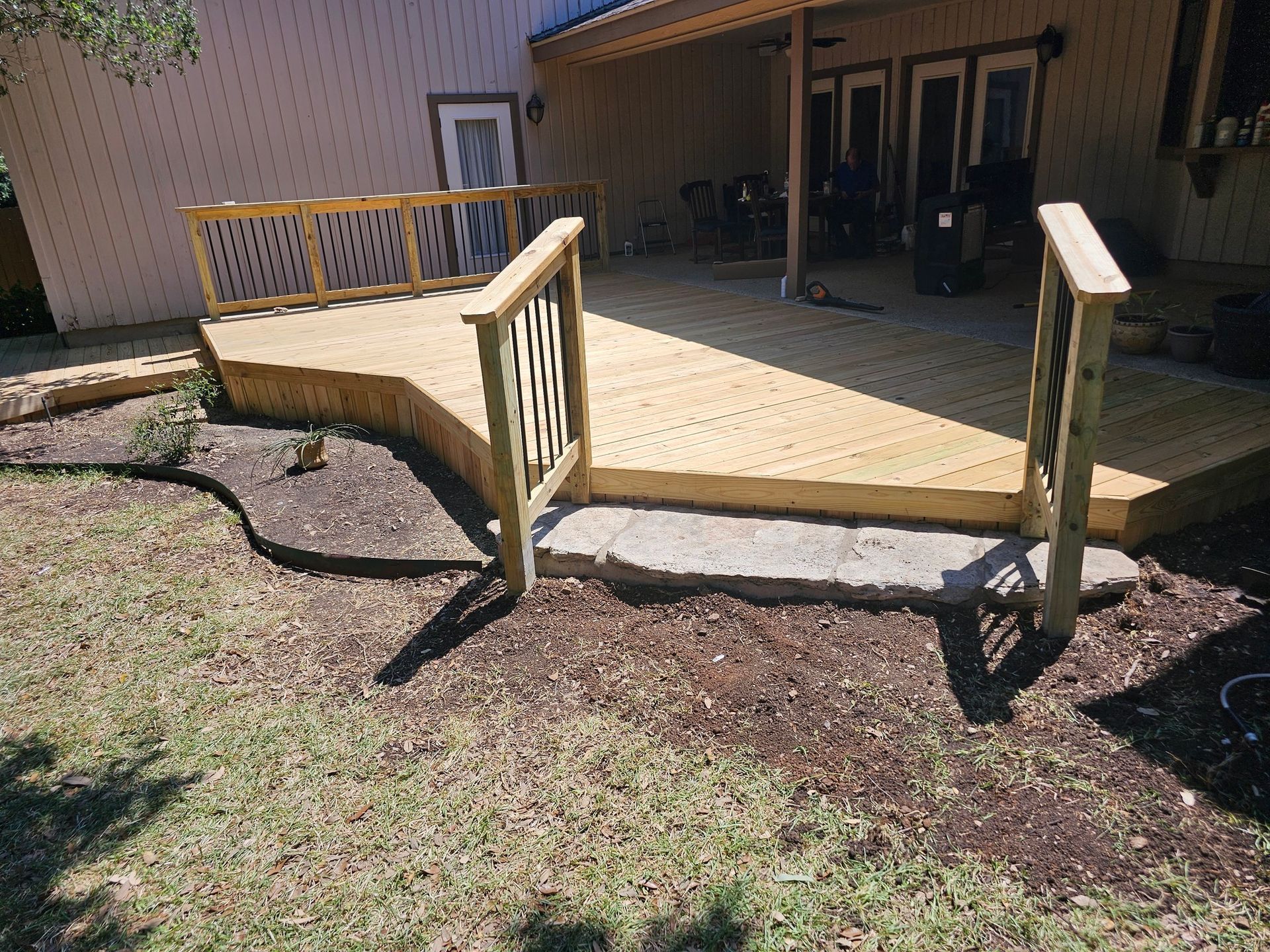 Newly constructed wooden deck with railing in a backyard. The deck is next to a house, with a small garden bed in front.