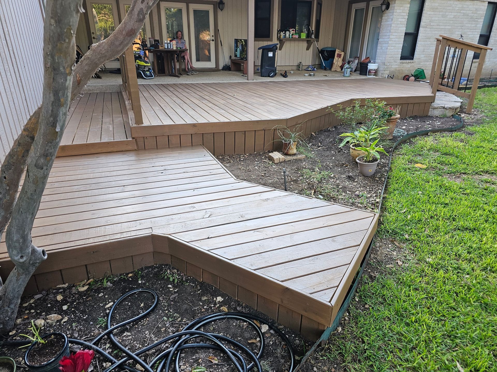 A multi-level composite deck with brown trim and tan decking borders a yard with grass and a tree.