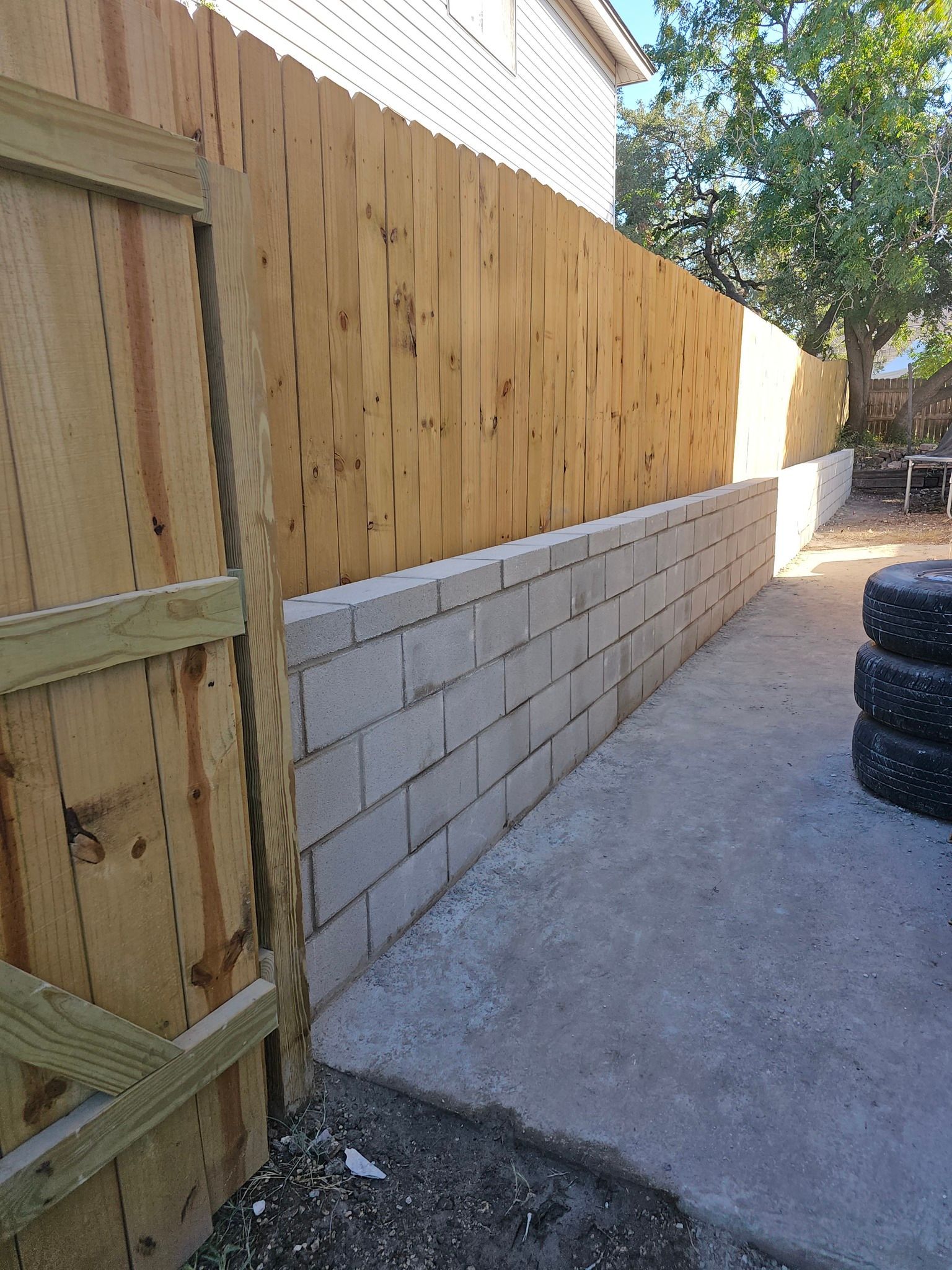 Wooden fence atop a cinder block wall. A gate is on the left; tires are stacked on the right. Concrete patio.
