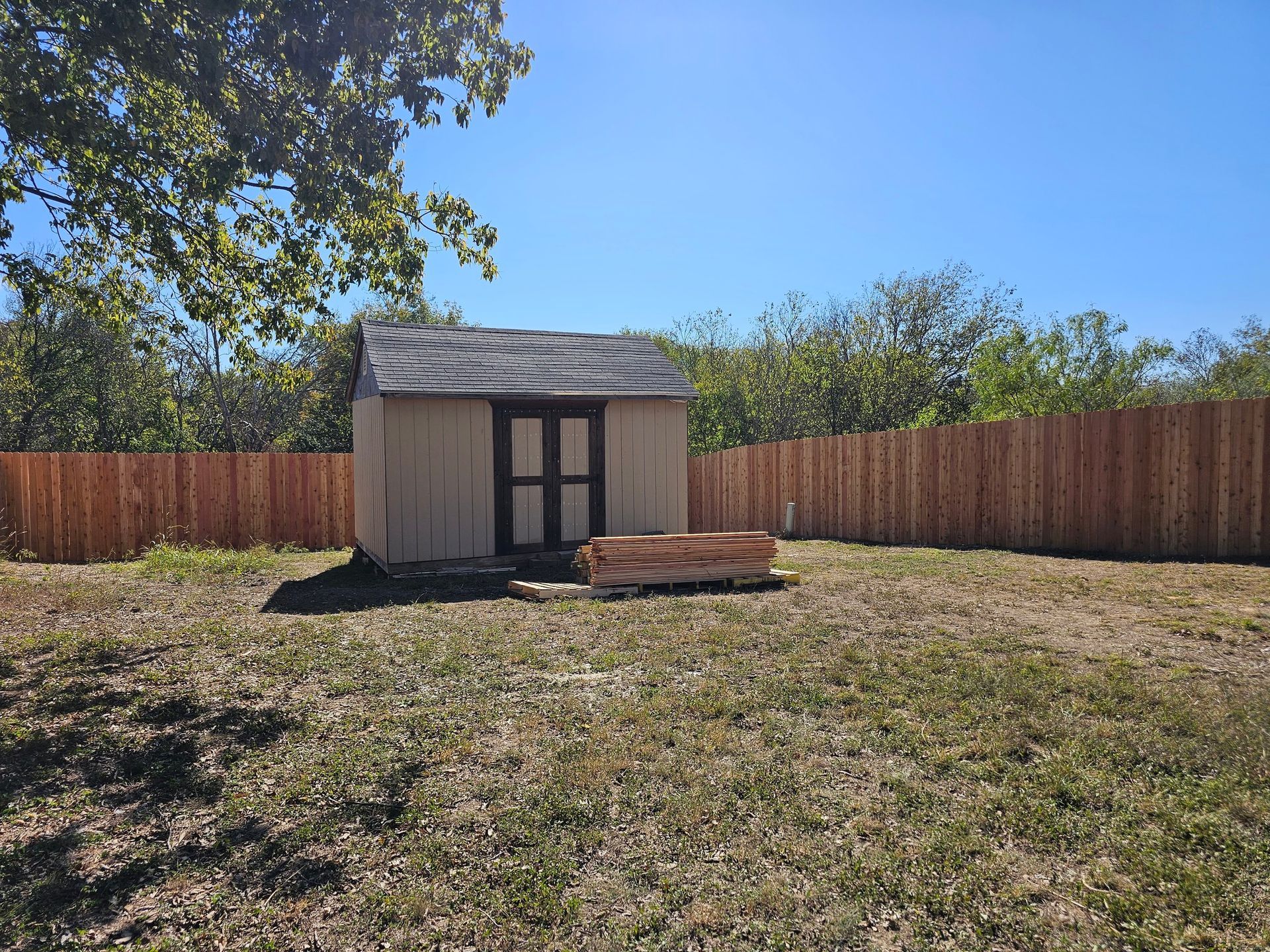 A tan shed with a dark roof and double doors sits in a grassy yard, bordered by a wood fence, under a bright blue sky.