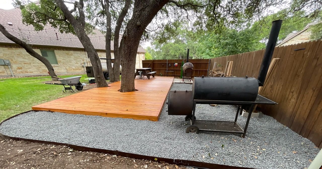 A backyard with a wooden deck built around a tree, a barbecue smoker on gravel, and a wooden fence.