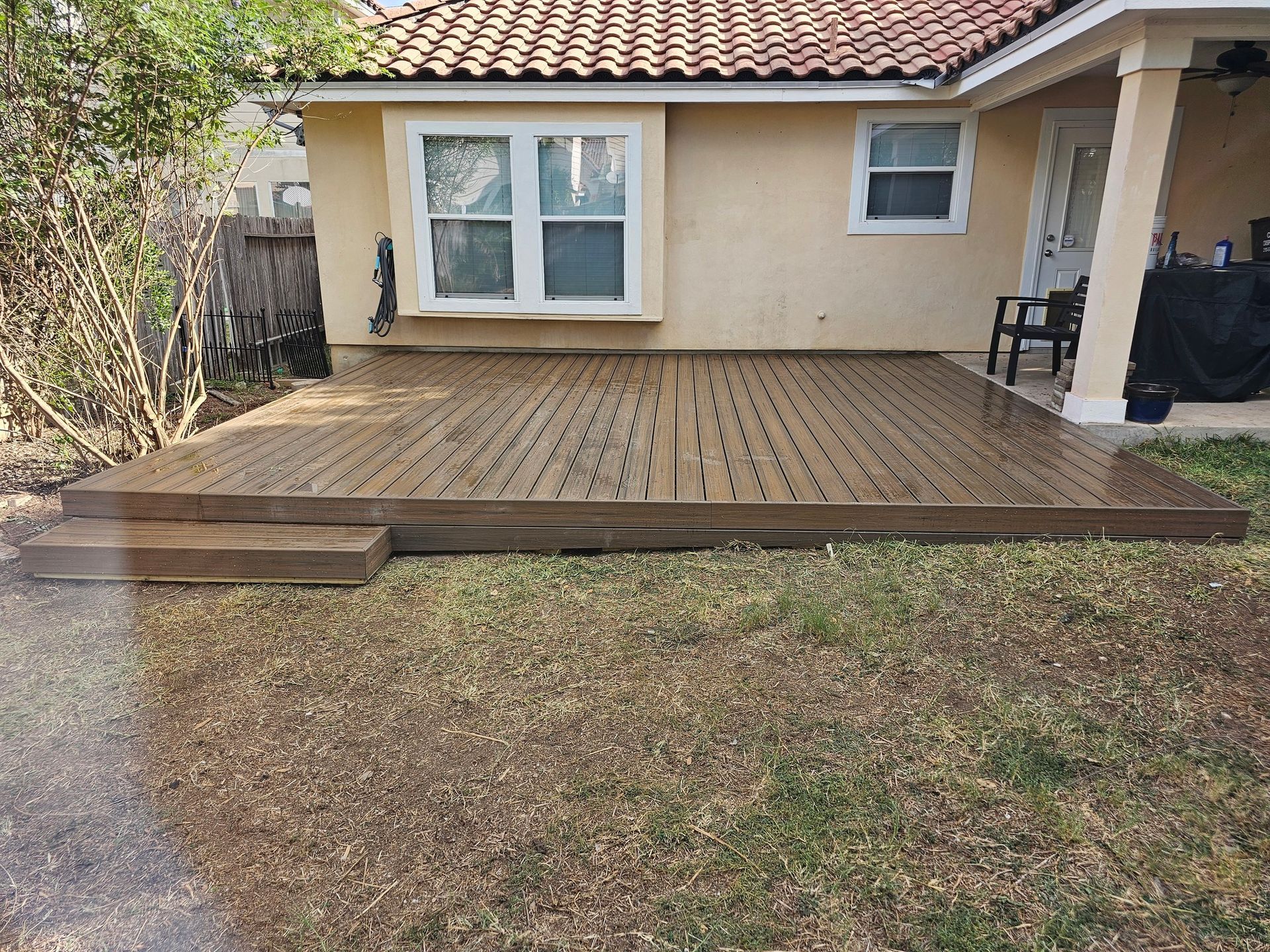 Newly built wooden deck with two steps, beside a tan stucco house with a window and doorway. The deck is on a grassy area.