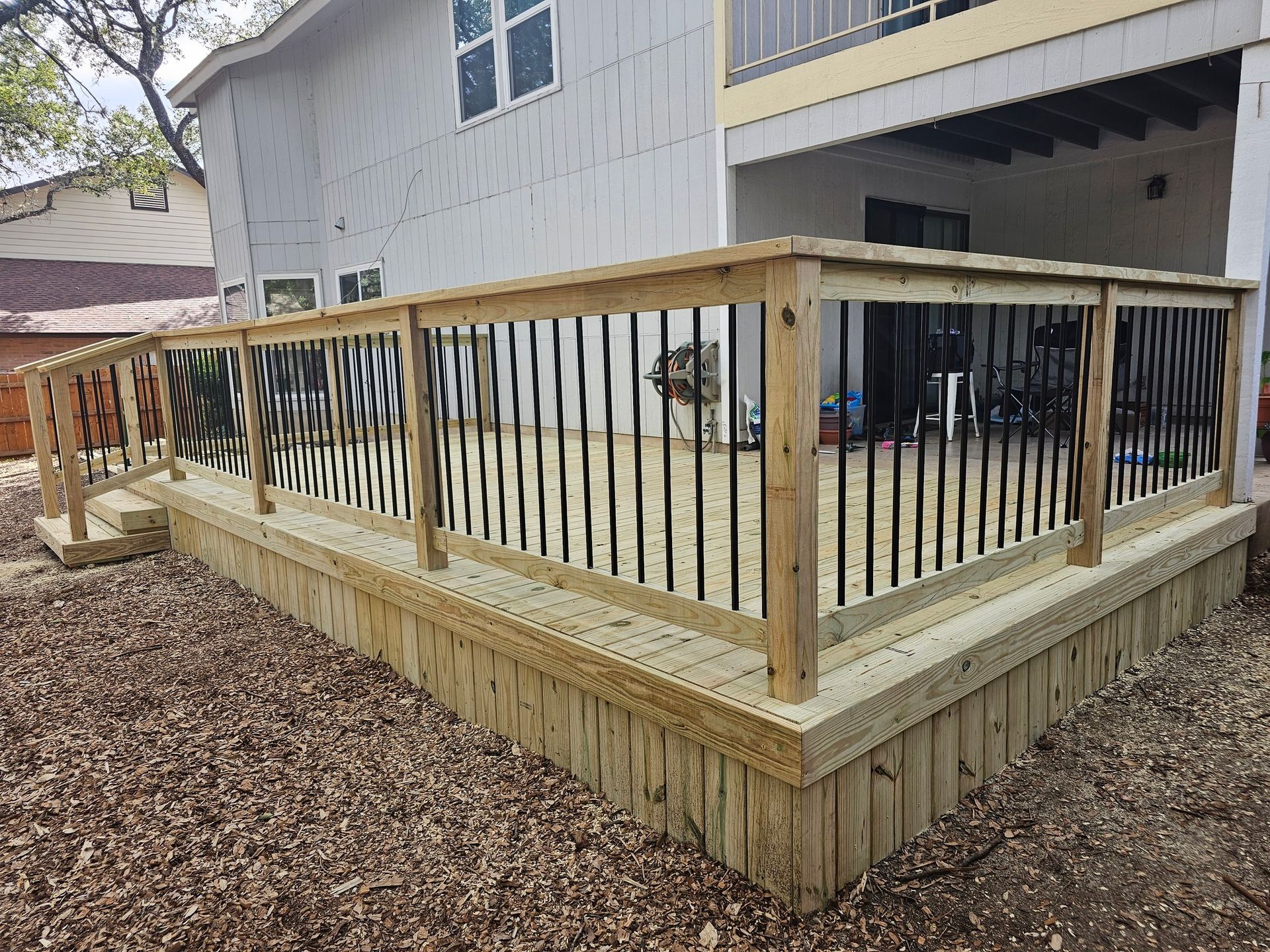 Wooden deck with black railing, built next to a house with beige siding, set on a bed of brown mulch.
