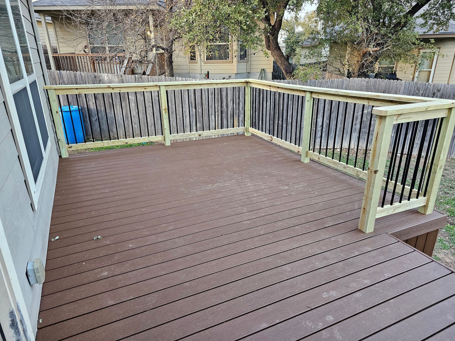 A brown composite deck with wooden railings and black vertical spindles, attached to the side of a beige house.
