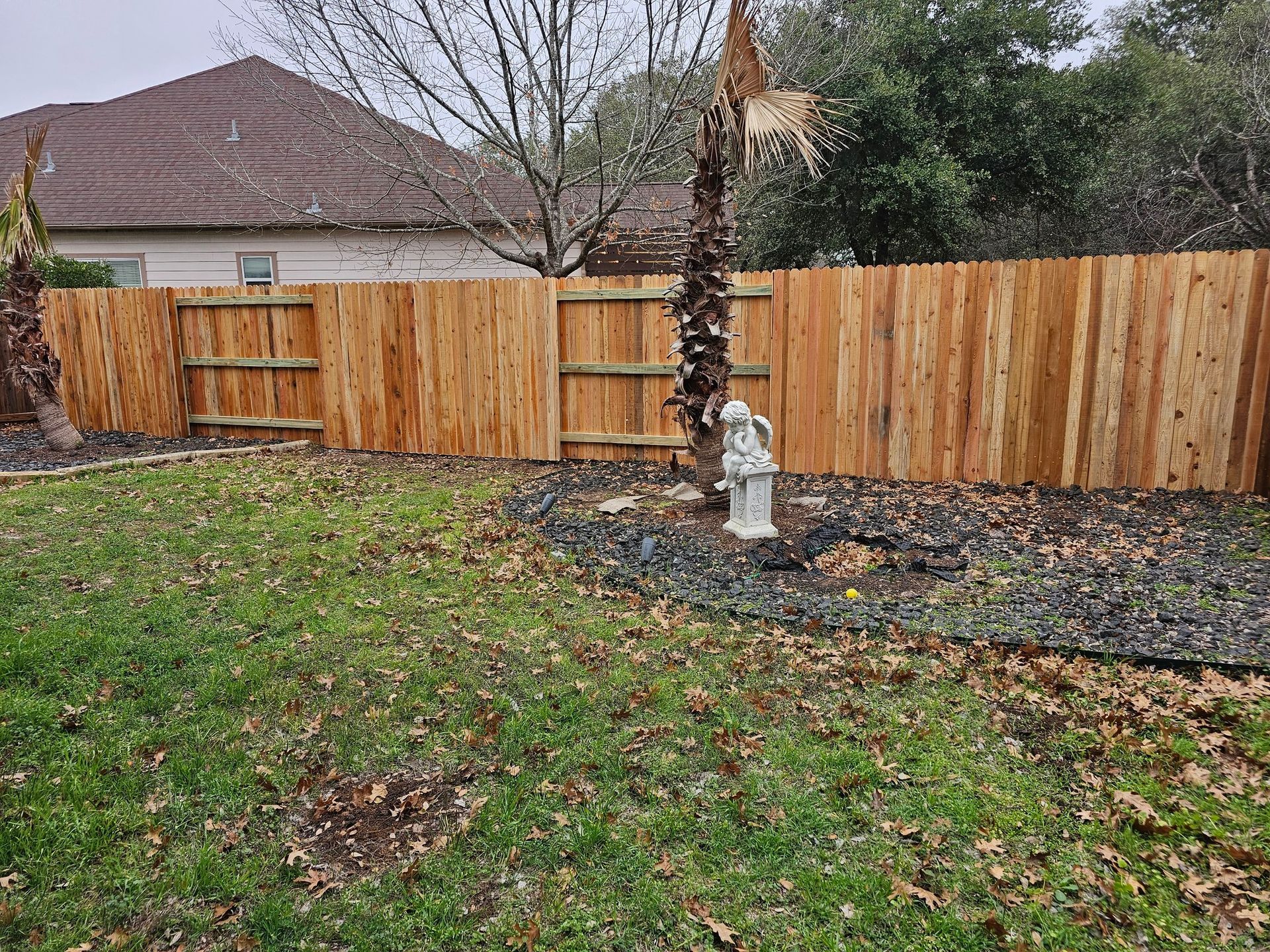Wooden fence bordering a backyard lawn, a stone statue stands near a palm tree, with a house in the background.