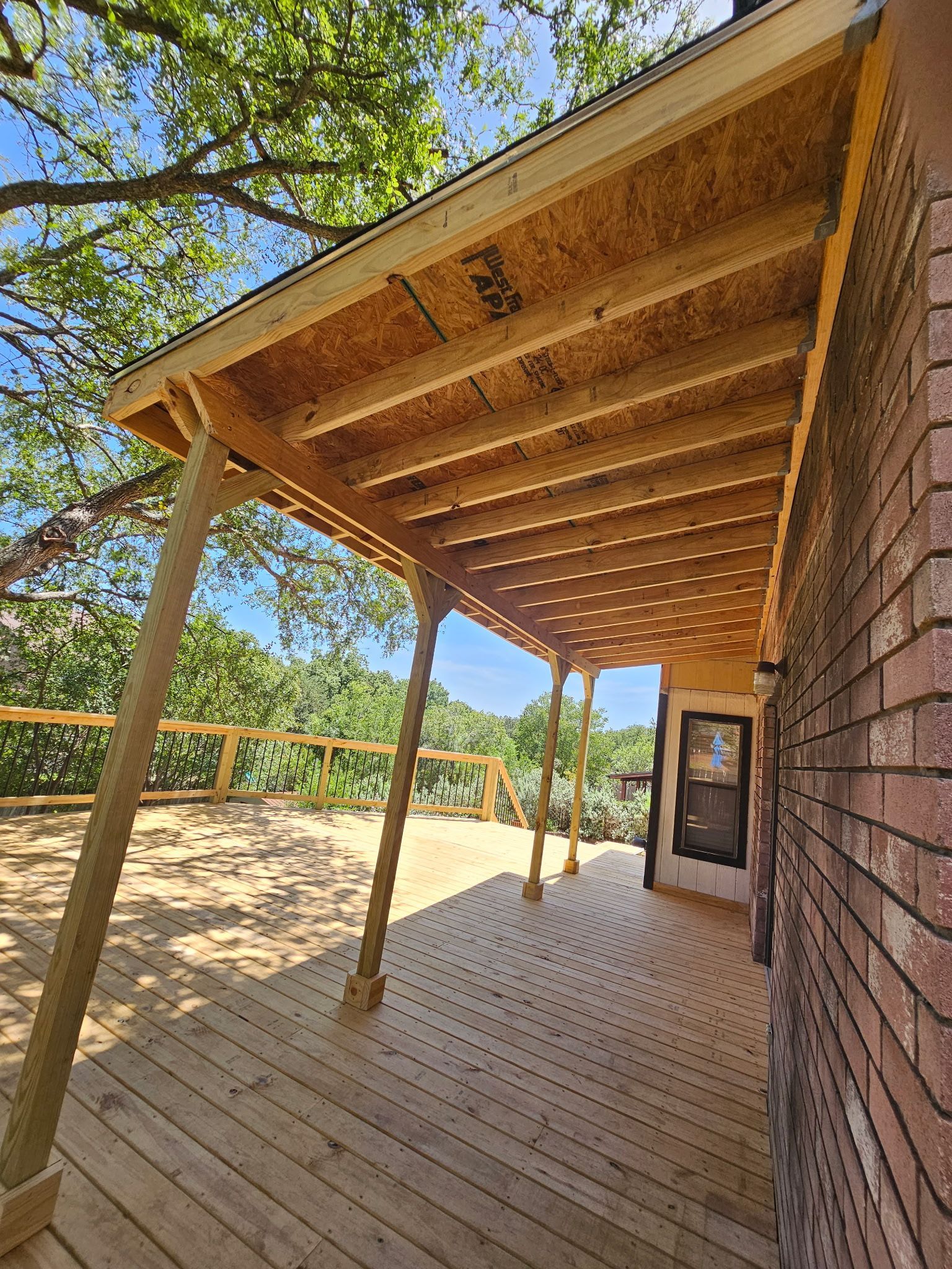 Wooden deck with attached roof supported by posts.  Brick house on right, trees and sky visible.