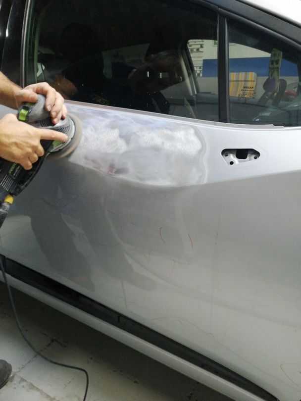 Una persona sta levigando con una levigatrice elettrica la portiera argentata di un'auto ammaccata in un garage.