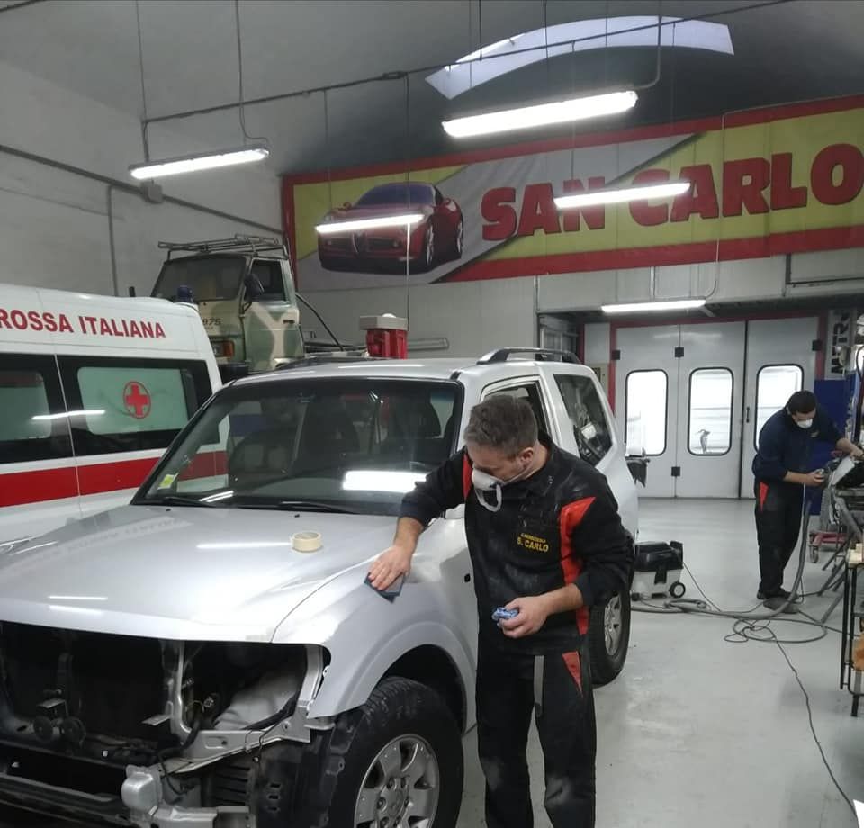 Un meccanico ispeziona un'auto bianca in un'officina, con l'insegna SAN CARLO sullo sfondo.