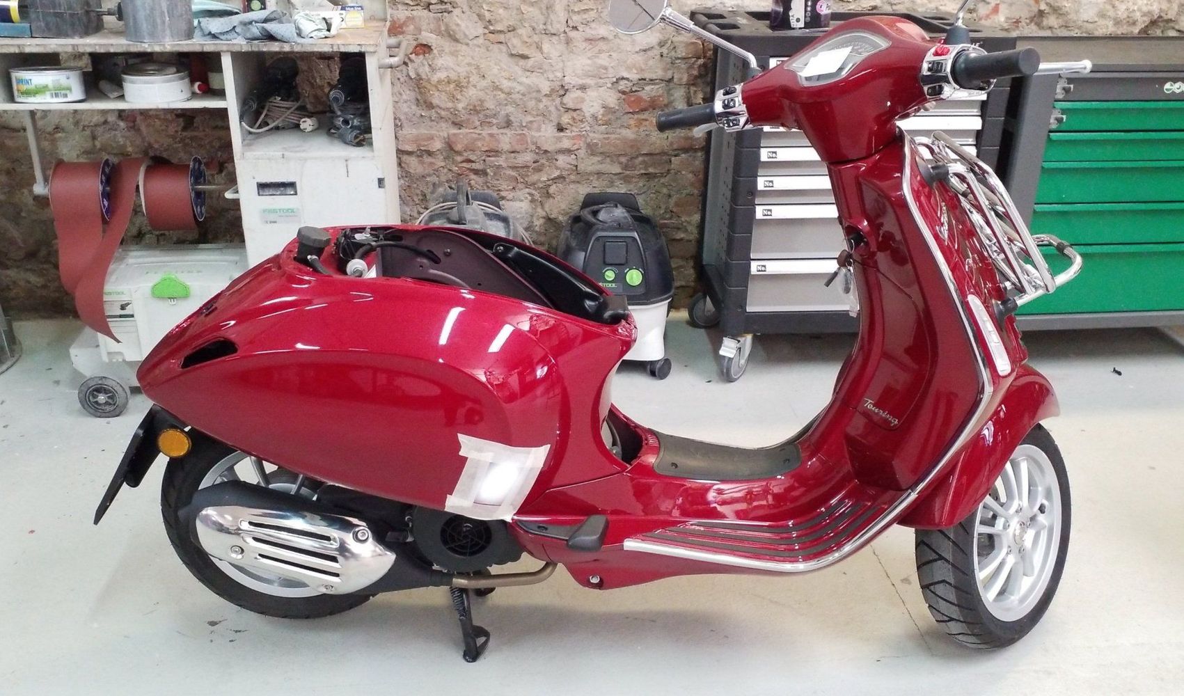 Uno scooter rosso in stile Vespa parcheggiato in un'officina, con attrezzi e attrezzature sullo sfondo.