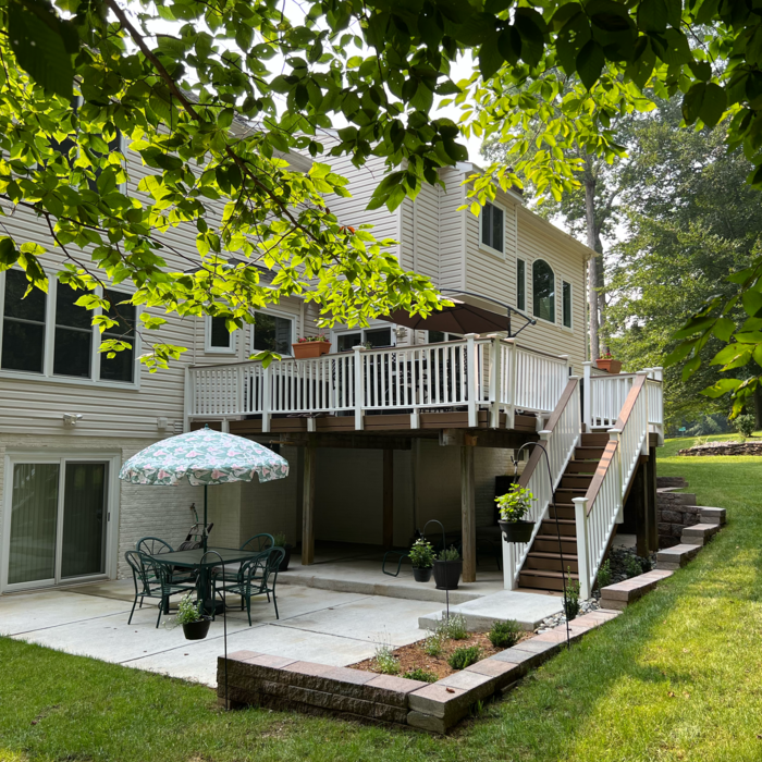 A two-story beige house with a deck and patio. A table and chairs sit under an umbrella on the patio.