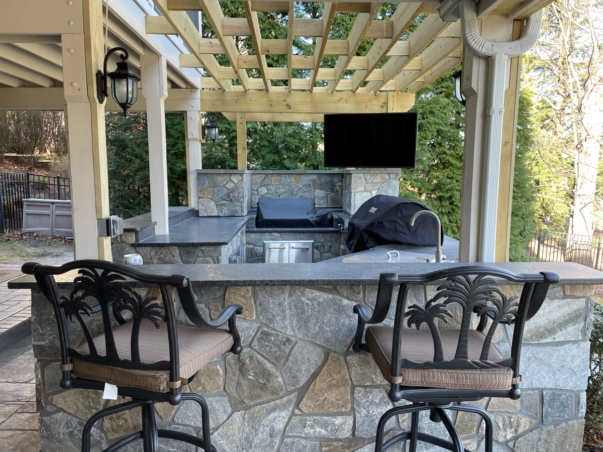 Outdoor kitchen with stone counters, grill, TV, bar seating, and wooden pergola.