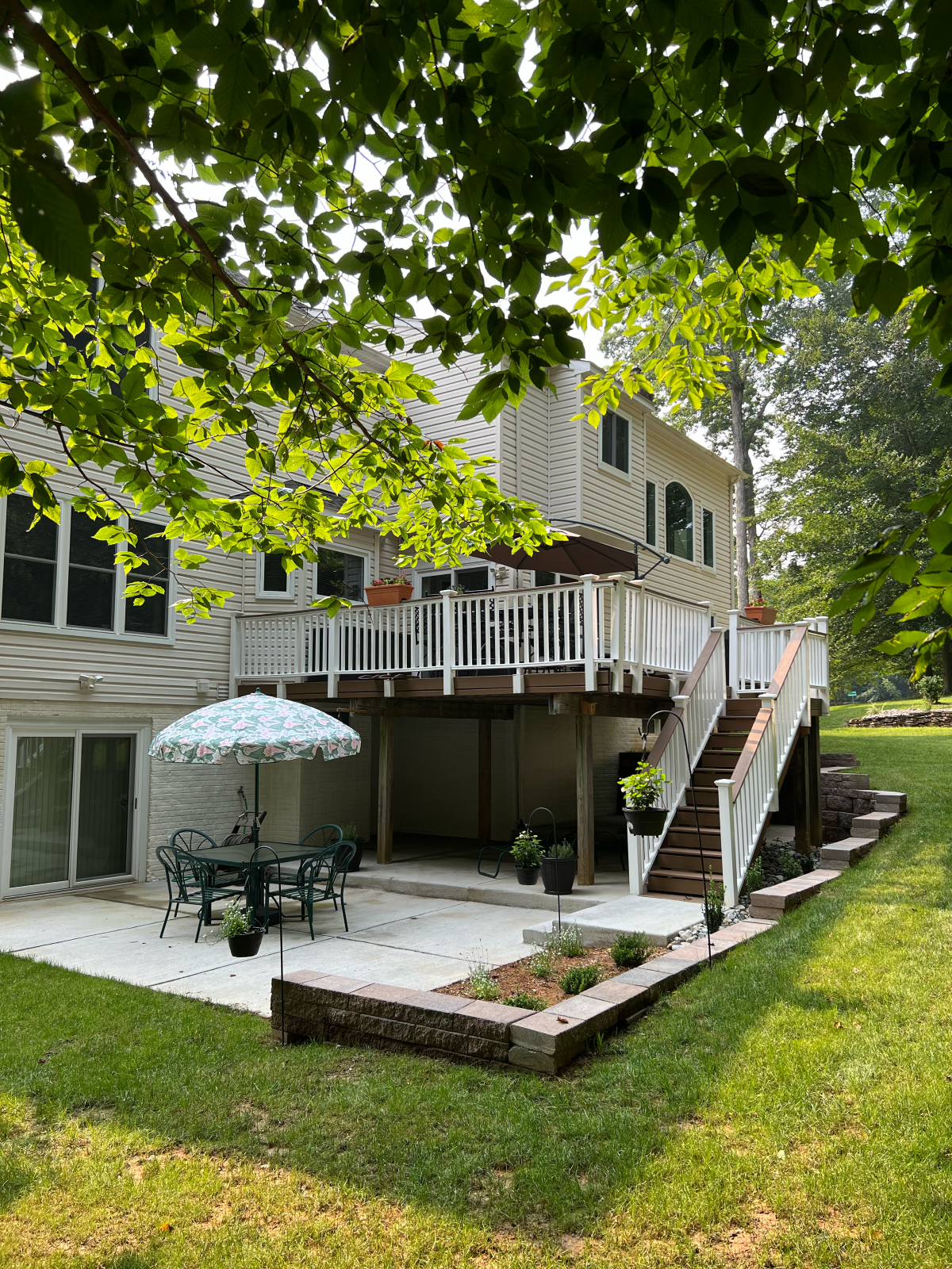Backyard patio with deck and stairs, green grass, and trees. White house with patio table and umbrella.