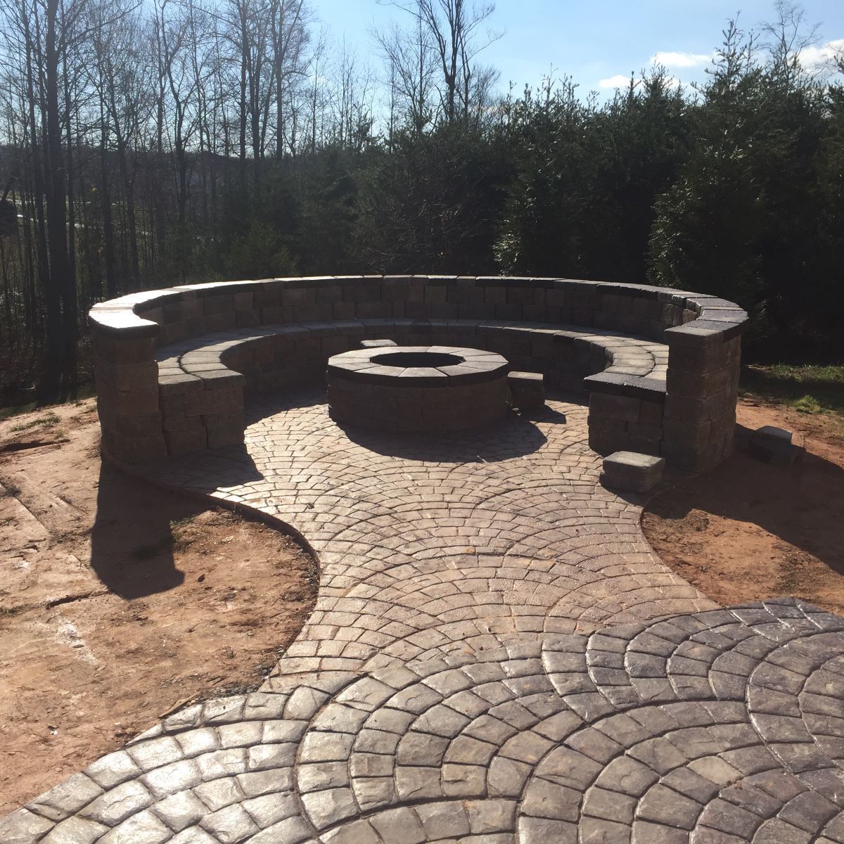 Circular stone fire pit with curved seating on a patterned brick patio in a yard with trees.