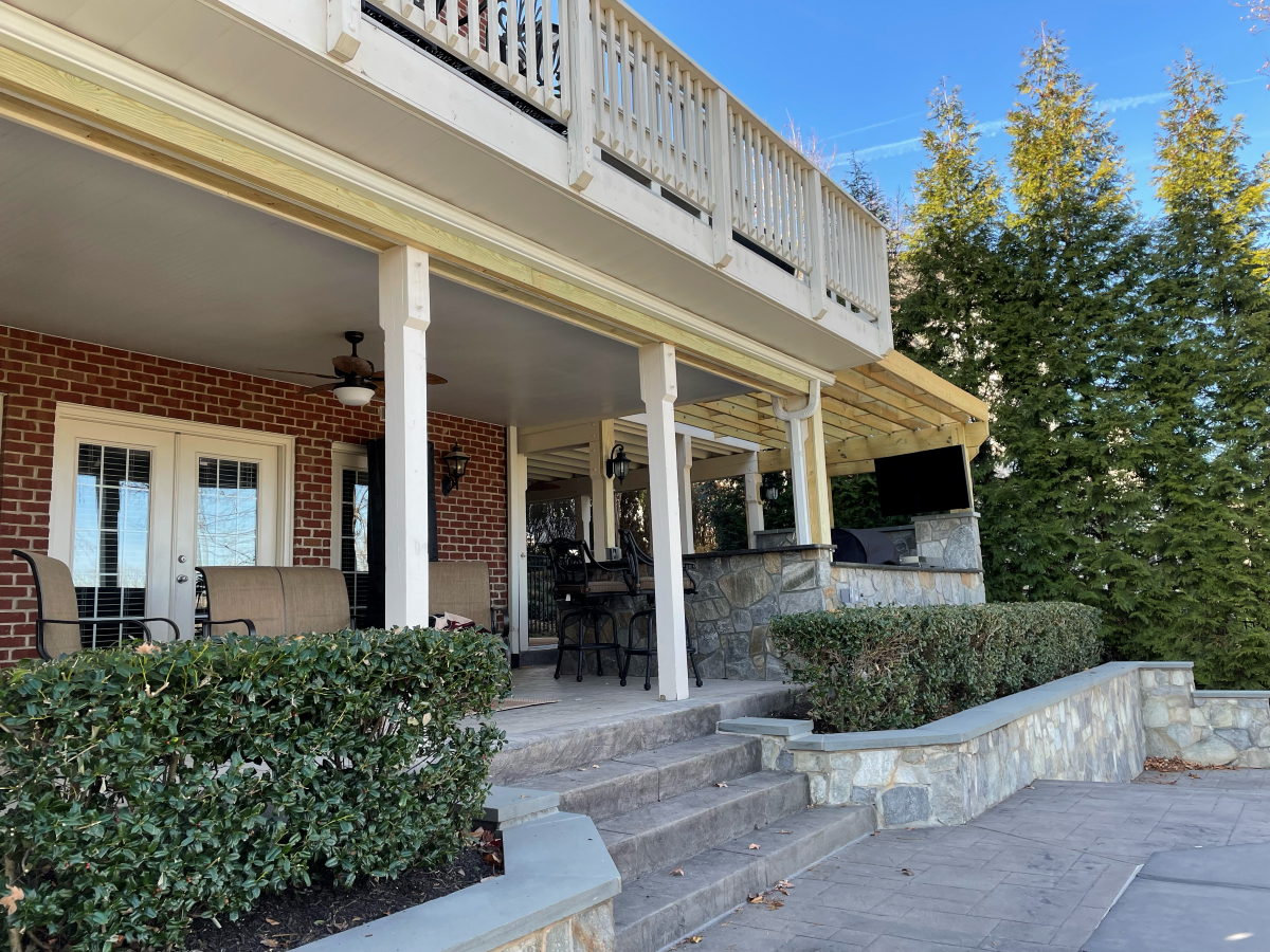 Brick house with a covered patio, stone steps, and a second-story balcony.  Green shrubs and a tree are also visible.