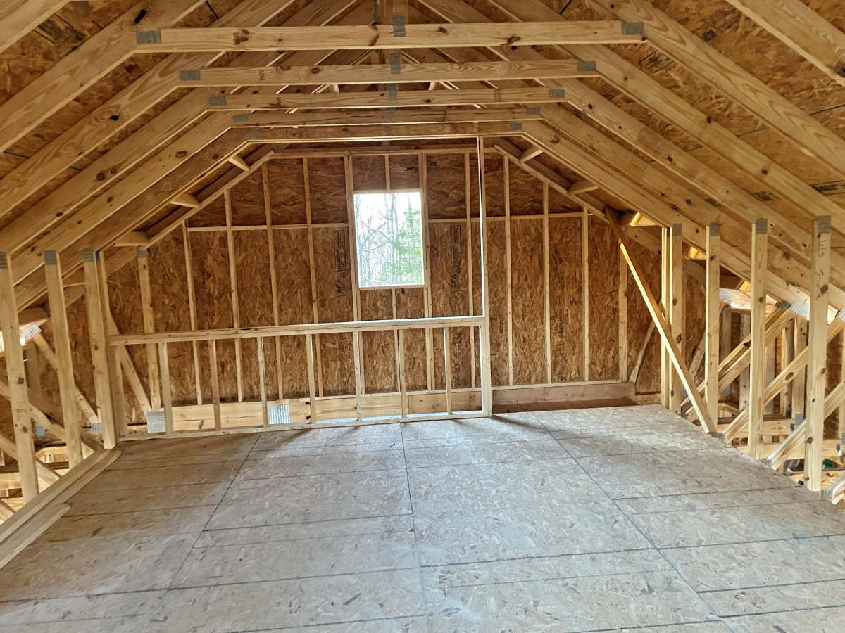 Interior view of an unfinished attic with wooden framing, sheathing, and a small window.