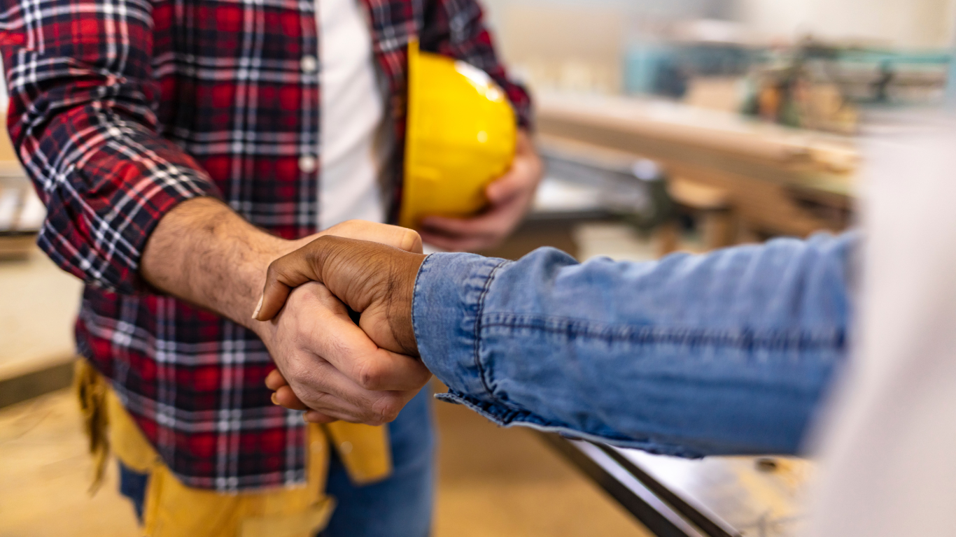 An electrical contractor wearing a hard hat is shaking hands with another man.