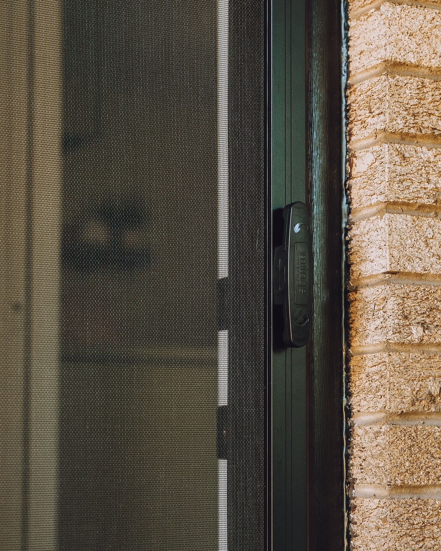 Close-up of a dark mesh screen door beside a tan brick wall and black handle