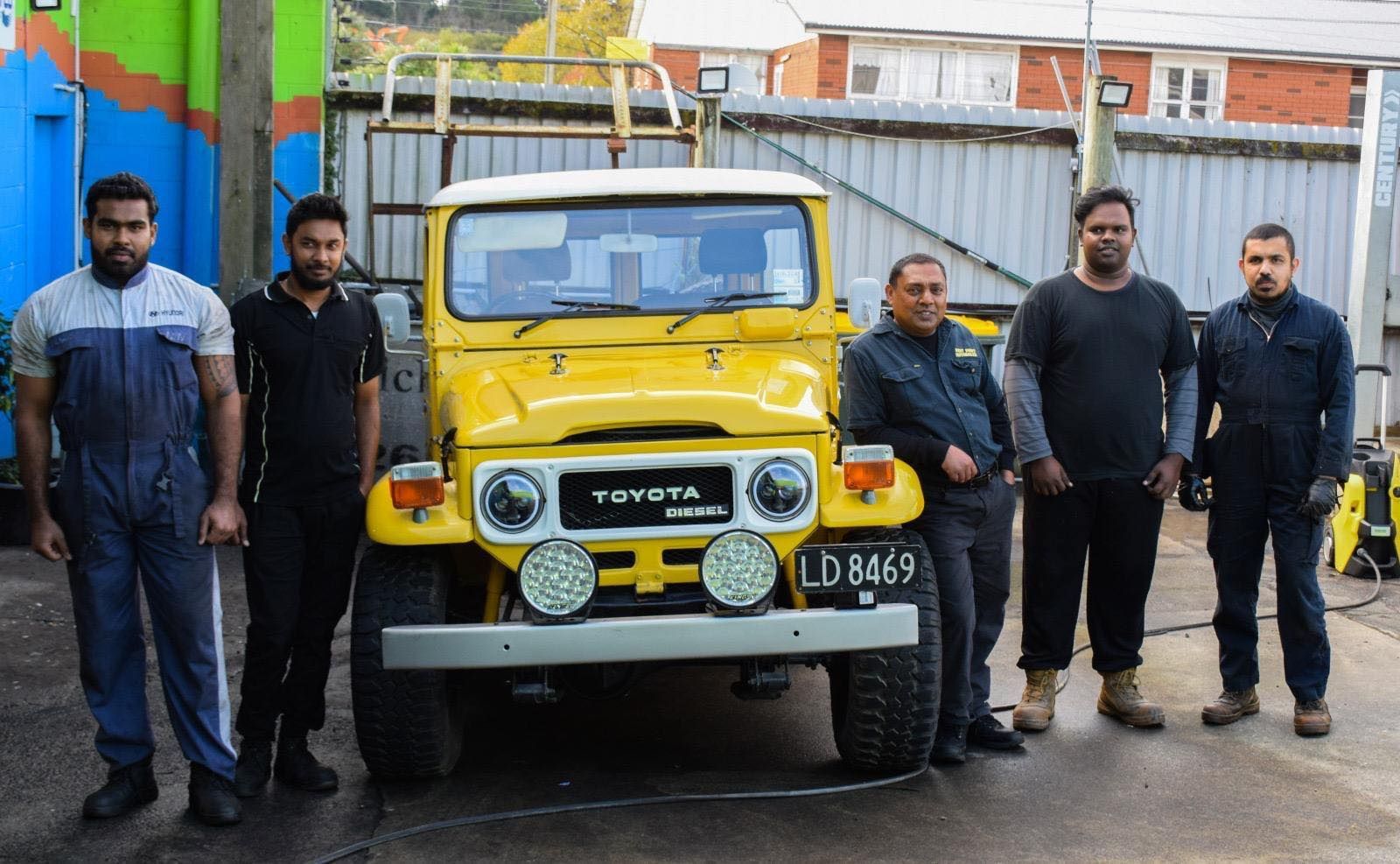Five men stand in front of a yellow Toyota Land Cruiser. They are outside a building with colorful walls.