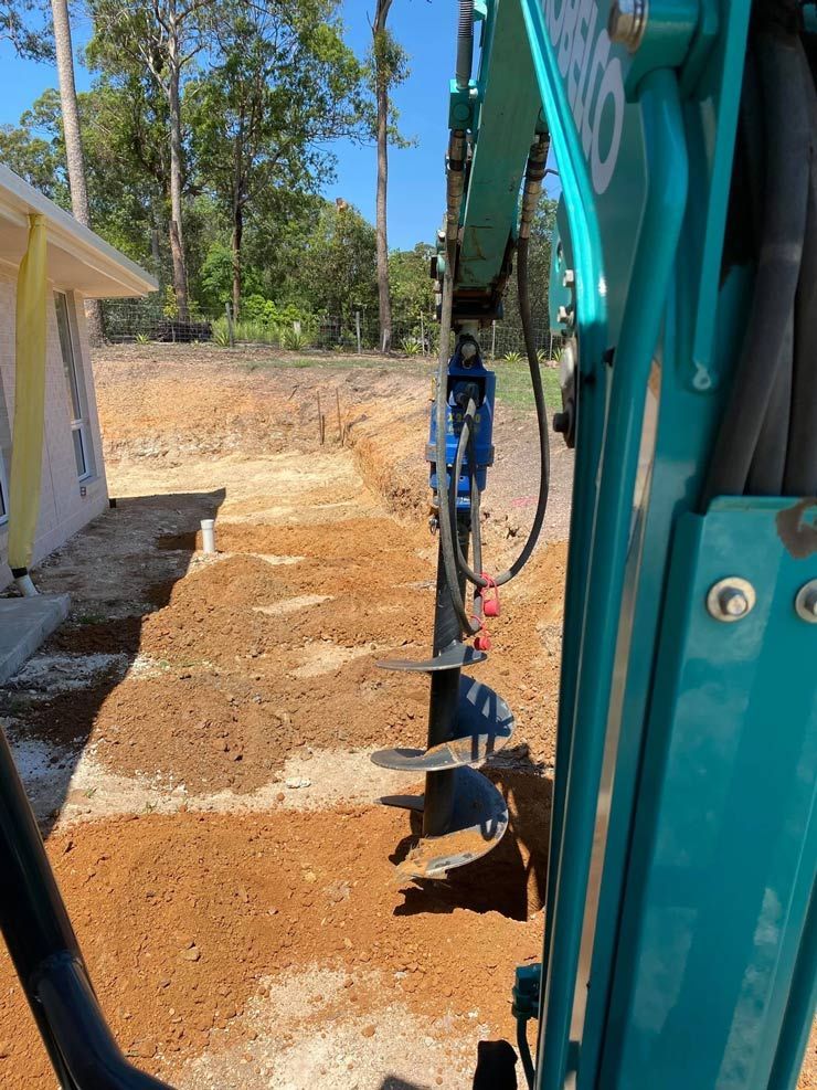 A Blue Excavator is Digging a Hole in the Dirt in Front of a House — Caloundra Landscape Supplies in Caloundra, QLD