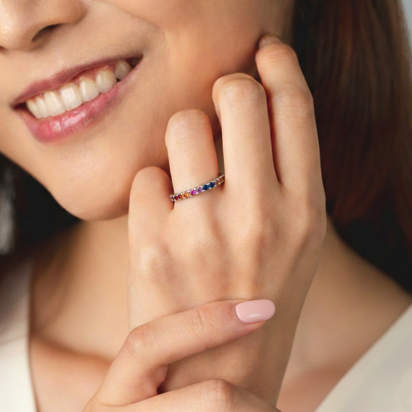 一位面帶微笑的人手指上戴著一枚纖細多彩的寶石永恆戒指,手輕輕放在臉旁。