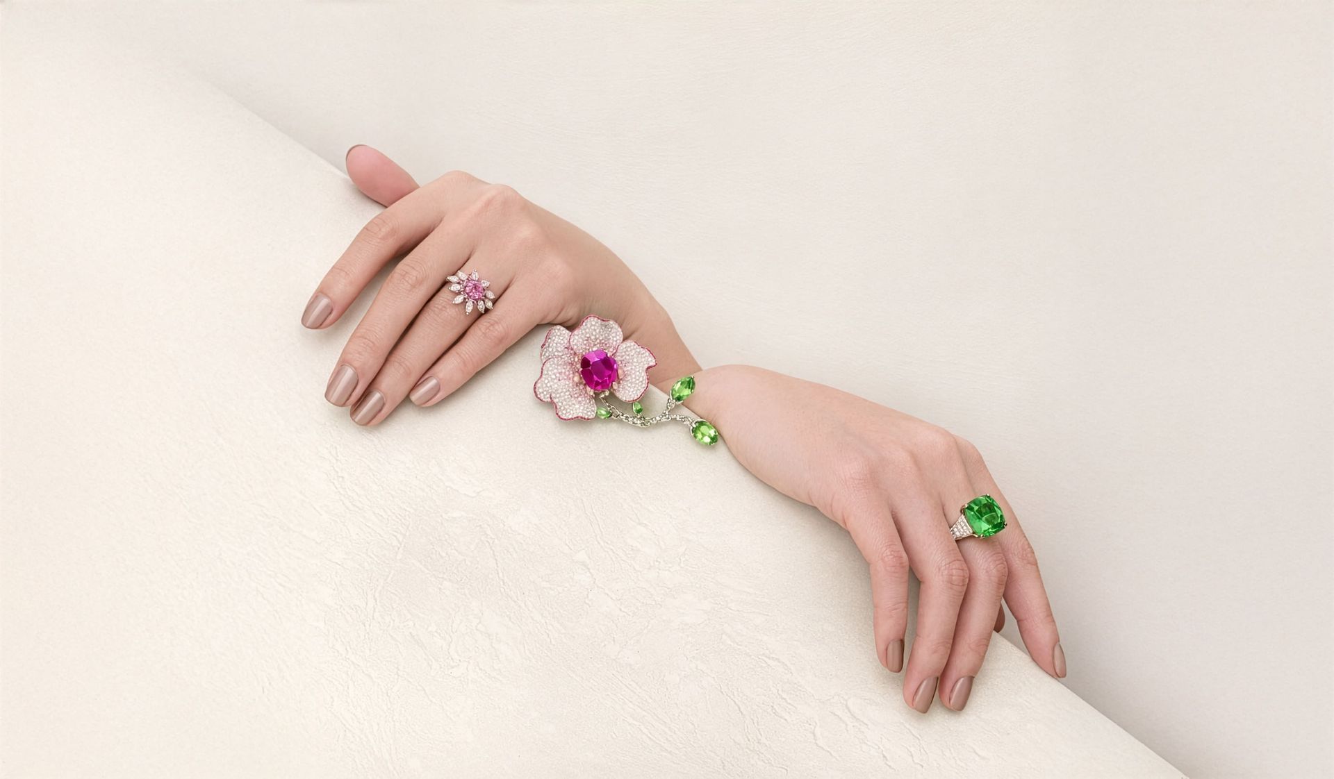 一雙修剪整齊的指甲,戴著花朵胸針、粉紅色寶石戒指和綠色寶石戒指,背景是白色。
