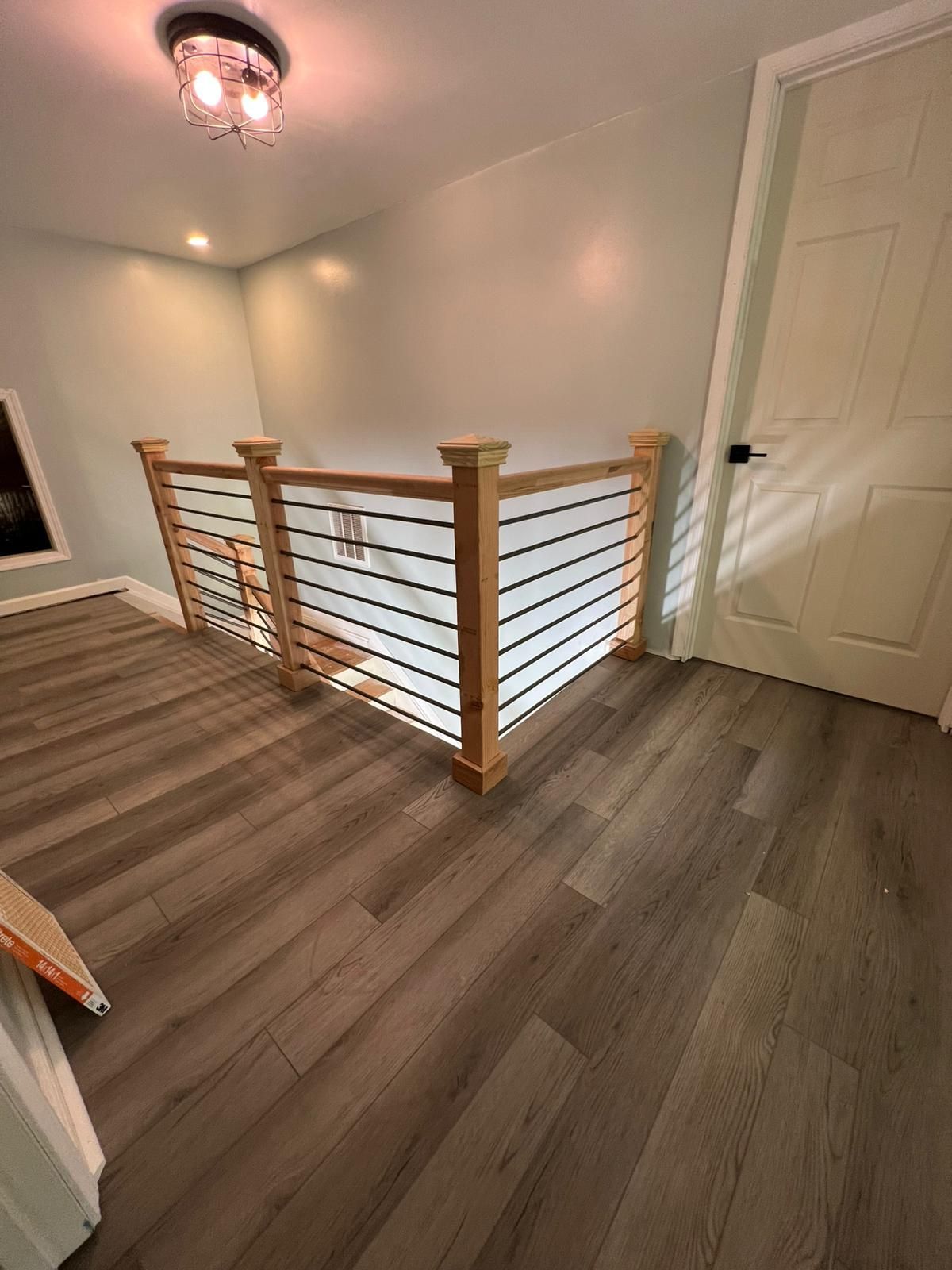 Stair landing with wood railing, gray flooring, white walls, and a closed door under a ceiling light.