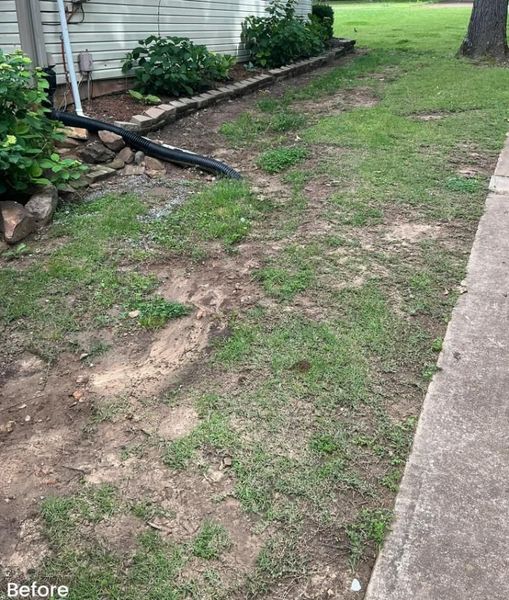 A yard with sparse grass, dirt patches, and a stone-edged flower bed alongside a house.