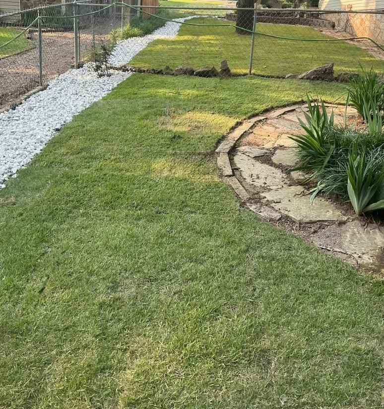 Lawn with a white rock-lined drainage path, a stone pathway, and a chain-link fence in a backyard.