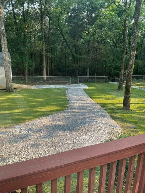 Gravel path leading through green grass towards a treeline and a fence, viewed from a wooden deck.