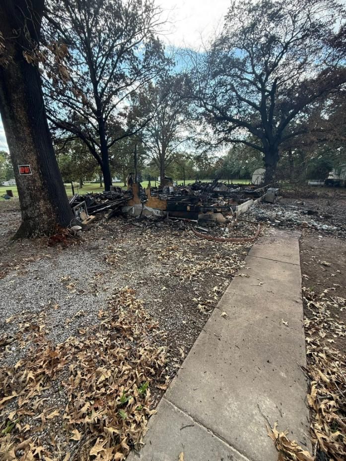 Charred remains of a structure after a fire, concrete path in foreground, trees in background, overcast.