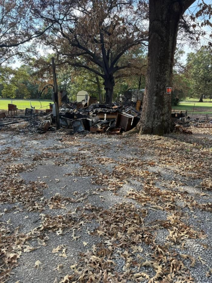 Charred remains of a structure. A large tree stands next to the debris on a surface covered in leaves.