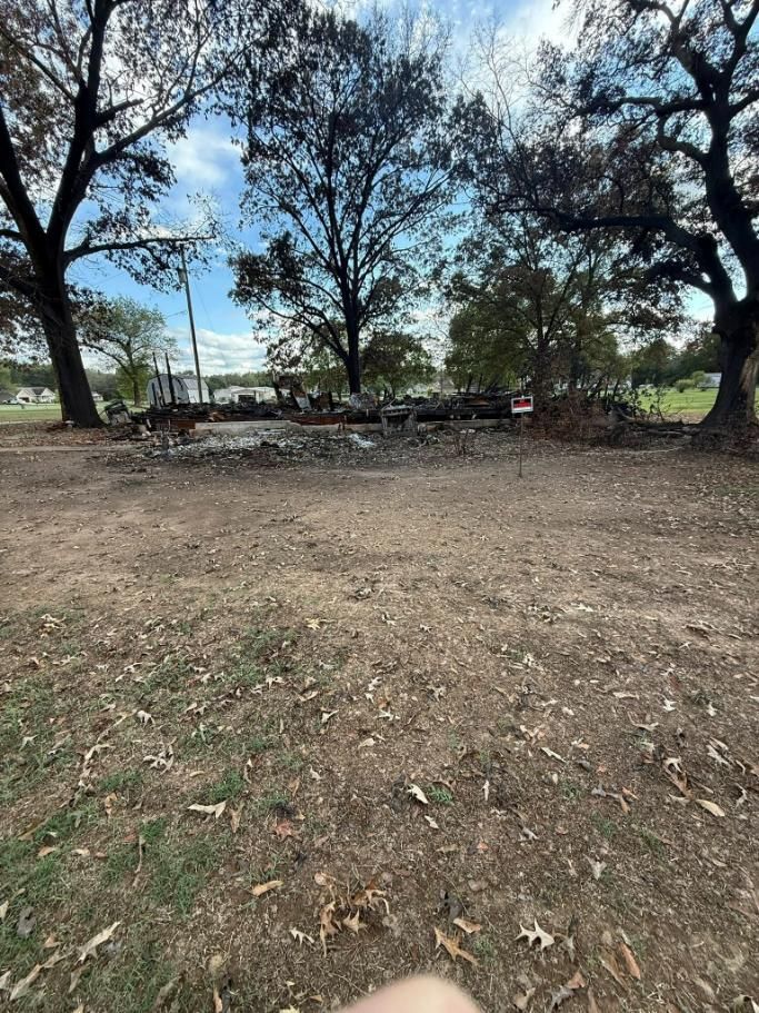 Debris and remnants of a structure amid a dirt field, with trees in the background under a cloudy sky.