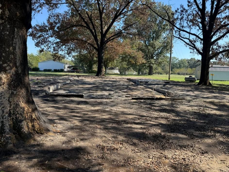 Dirt lot with wooden beams, trees, and houses in the background on a sunny day.