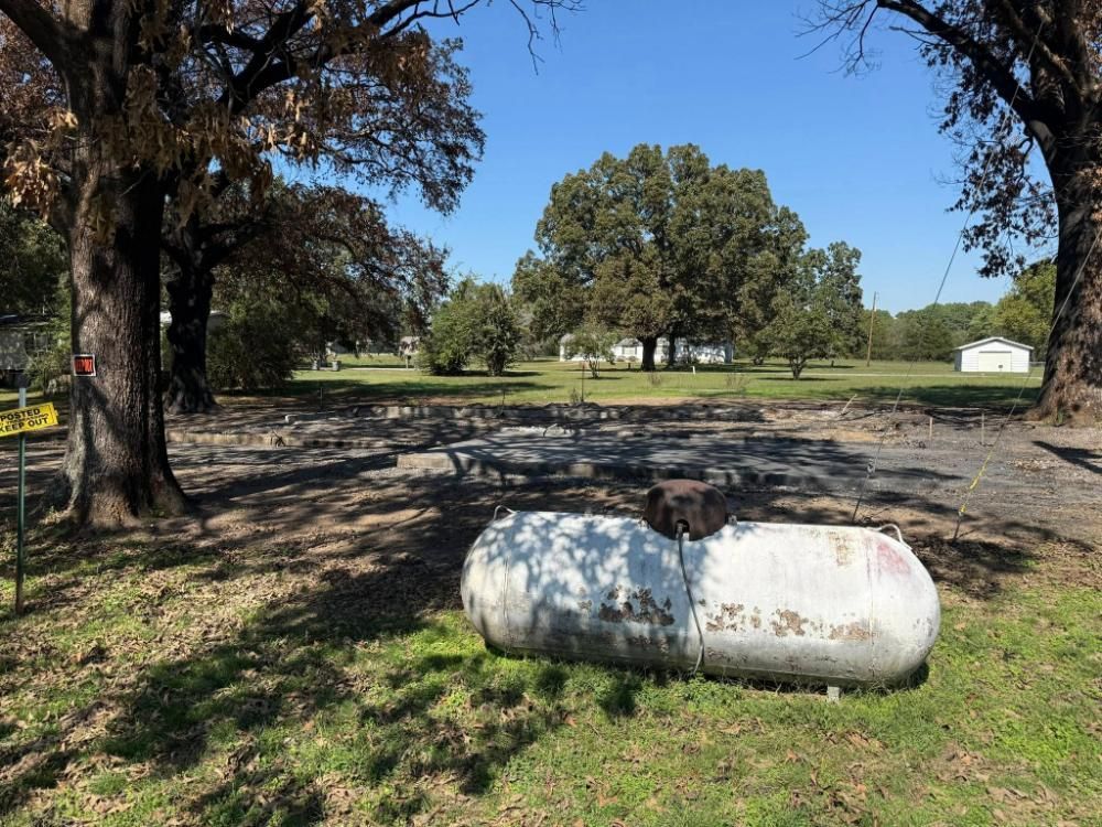 A large, weathered propane tank sits in a grassy yard, near a charred area, with trees and a house in the background.