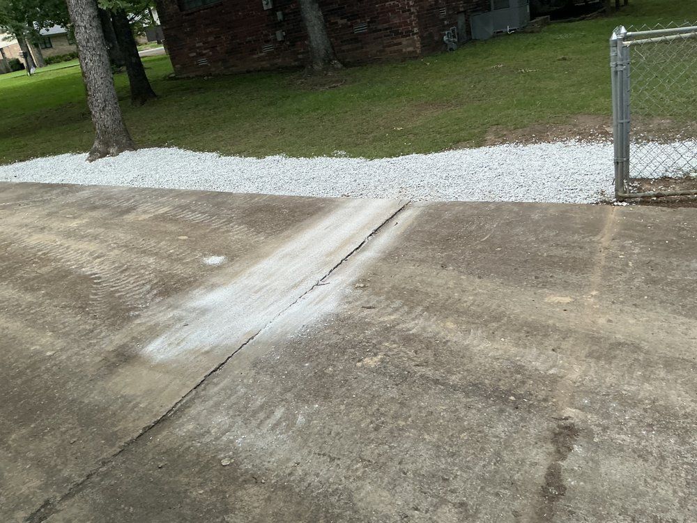 Concrete driveway with a crack, leading to a gravel border and a gate. Green grass and trees in the background.