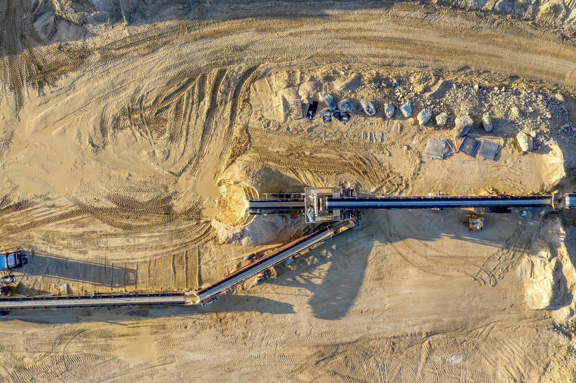 Aerial view of a quarry with conveyor belts transporting materials. Sandy-colored ground and machinery are visible.