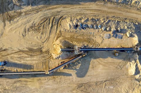Aerial view of a quarry with conveyor belts transporting materials. Sandy-colored ground and machinery are visible.