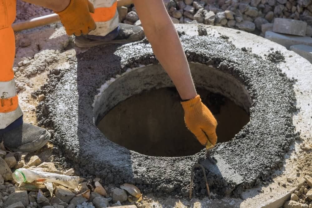 Person in orange gloves and work pants, smoothing concrete around a manhole.