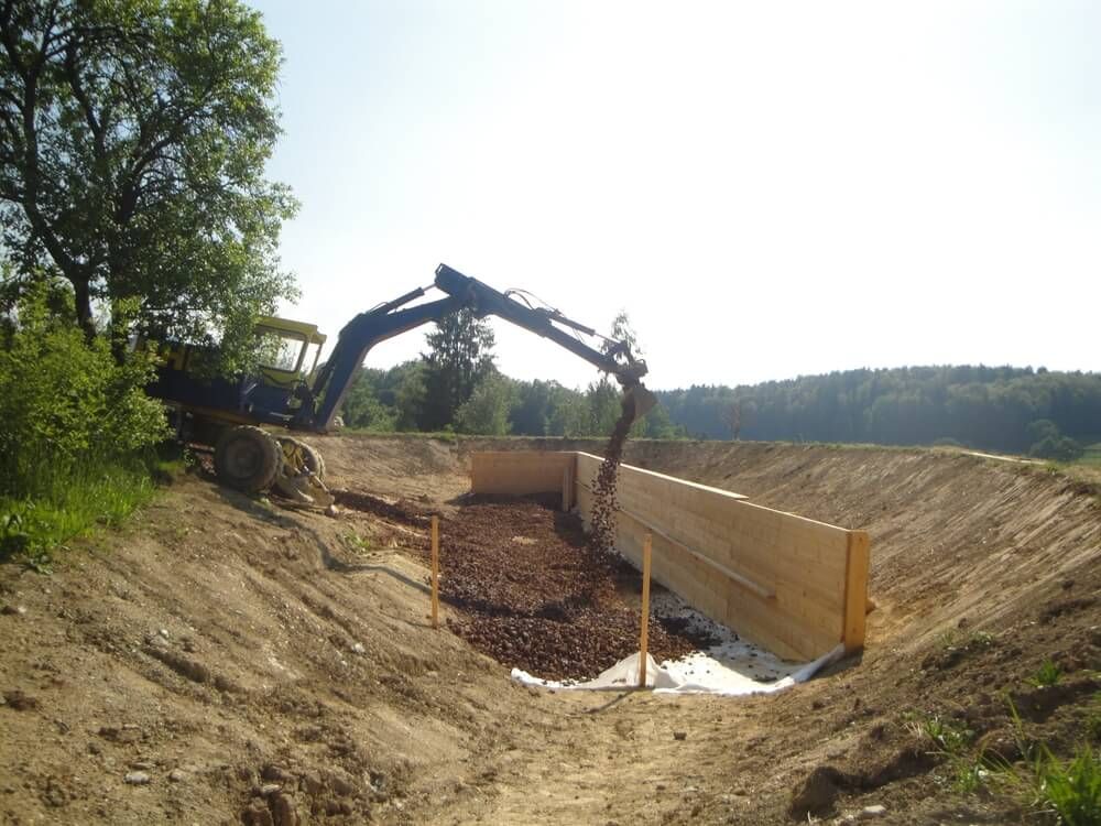 An excavator dumps soil into a trench lined with wood and fabric; outdoors on a sunny day.