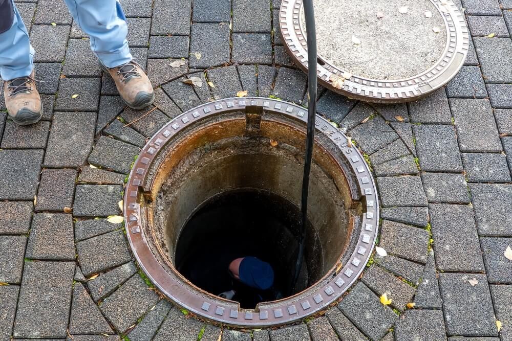 Person looking into open manhole. Brown brick pavement surrounds the opening and open cover.