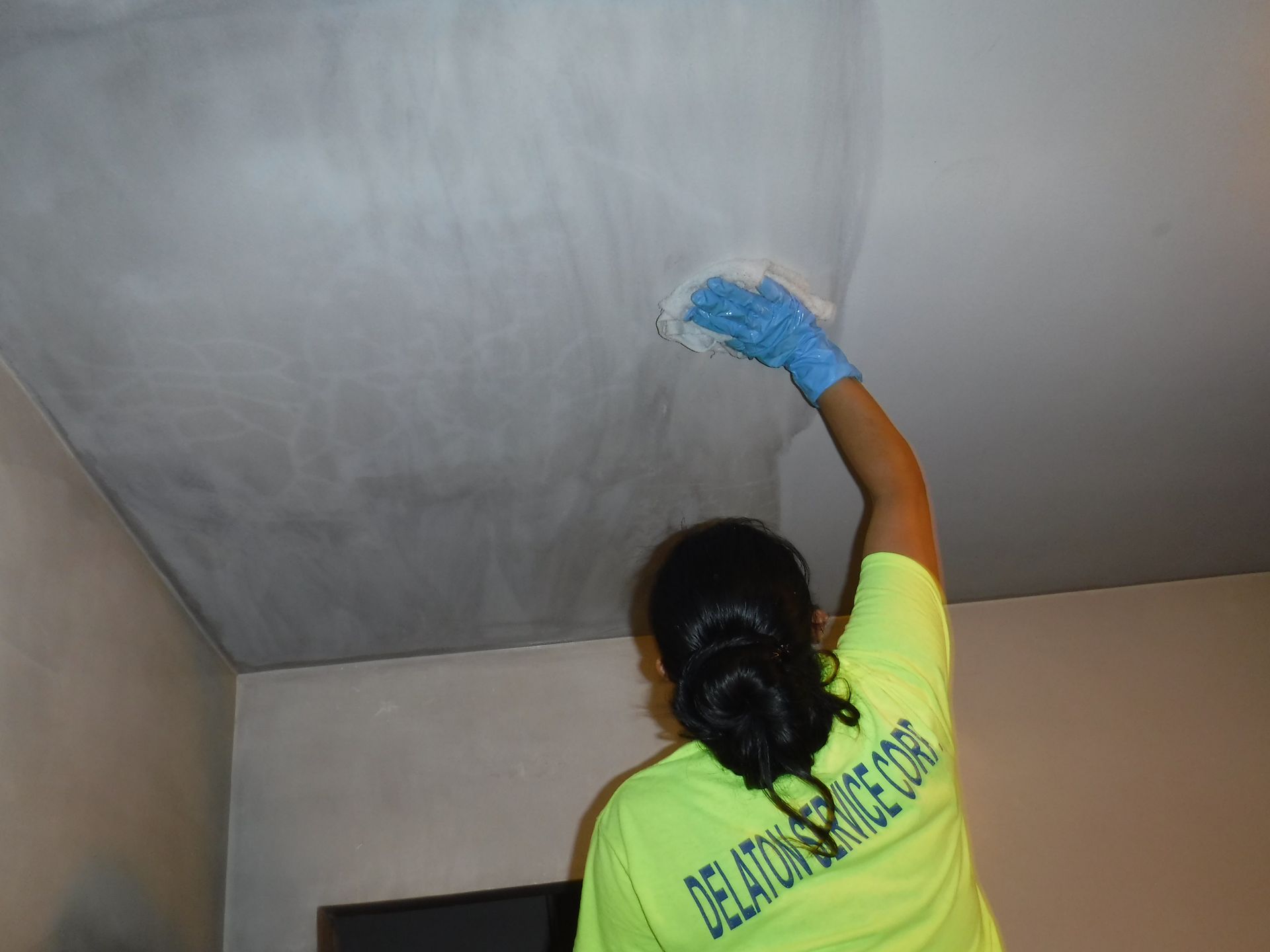 Person in yellow shirt and blue gloves wiping a light-colored ceiling.