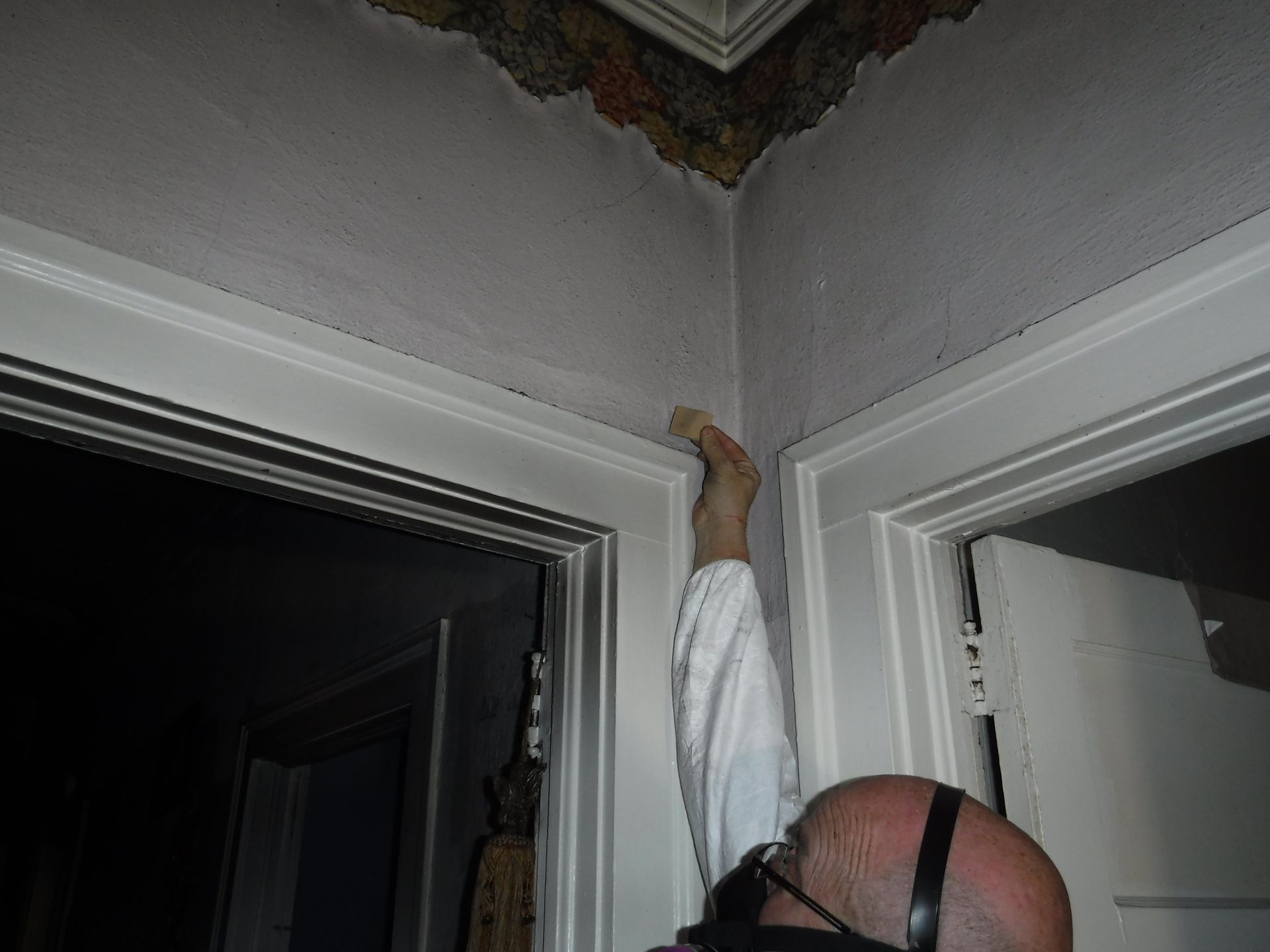 Person reaching up to damaged ceiling corner, white trim, doorways, and wearing a mask.