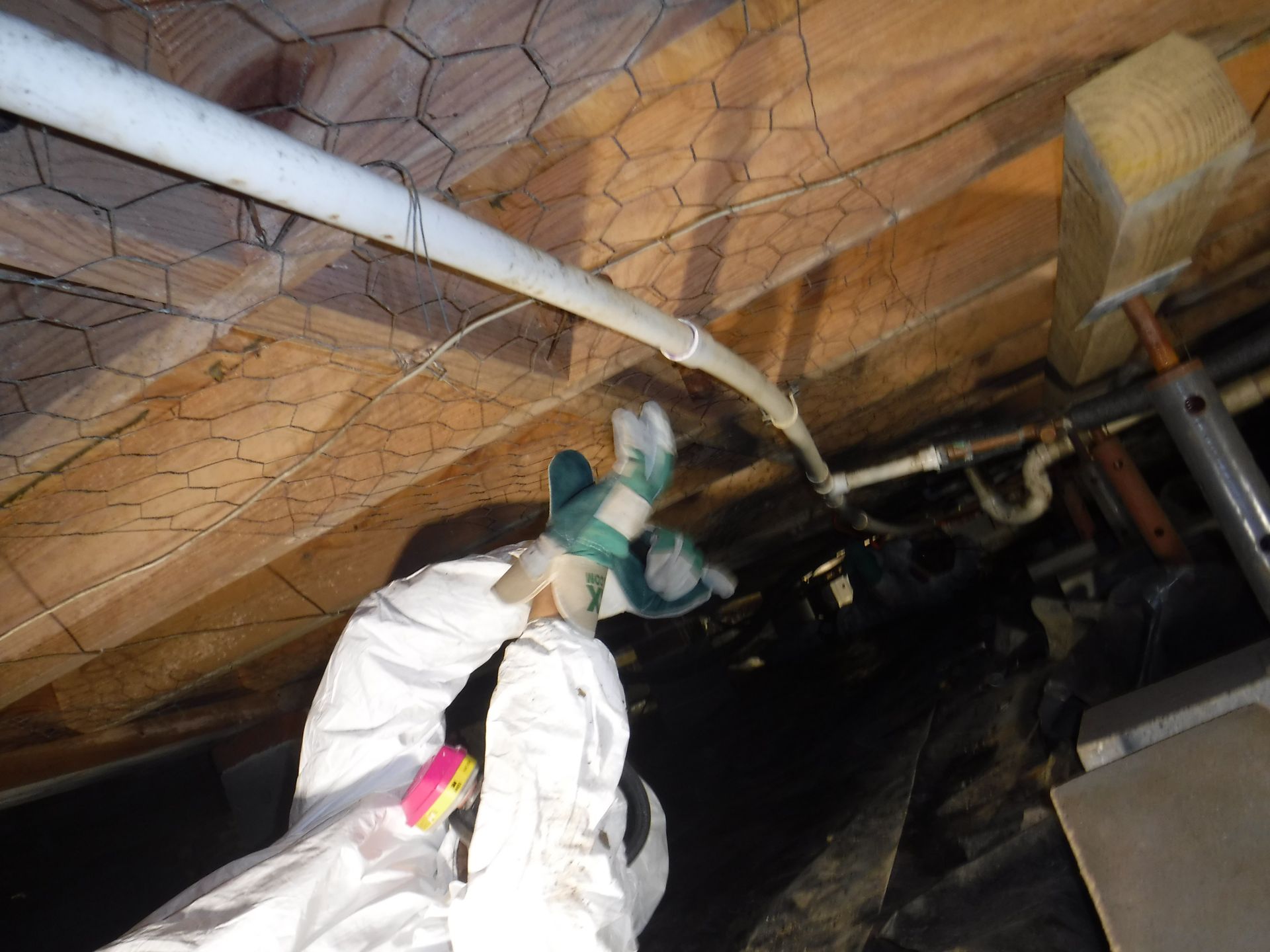 Person in protective suit working under a structure, near pipes and chicken wire.