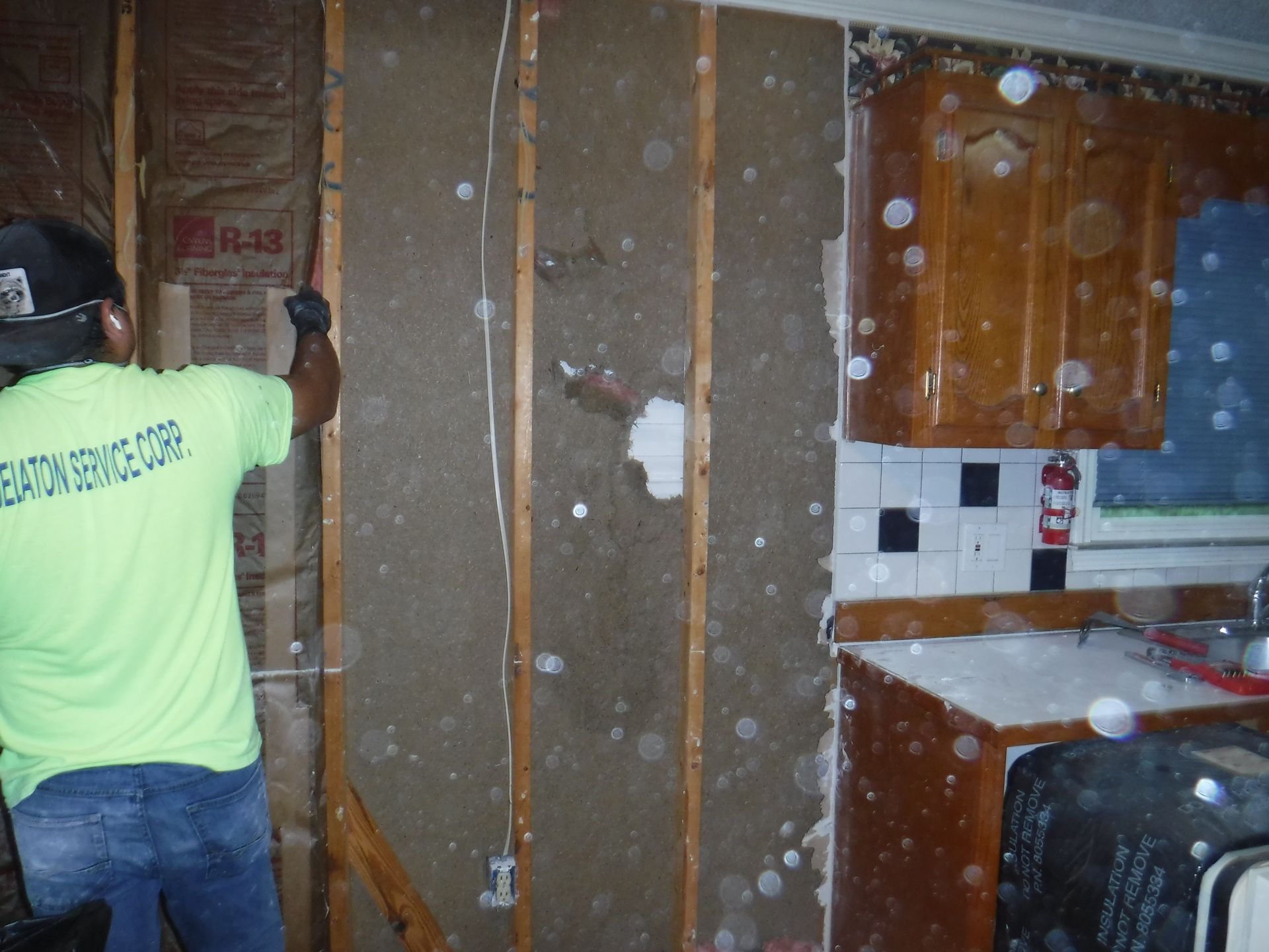 Man in neon shirt removing wallboard in a kitchen with water damage; insulation exposed.