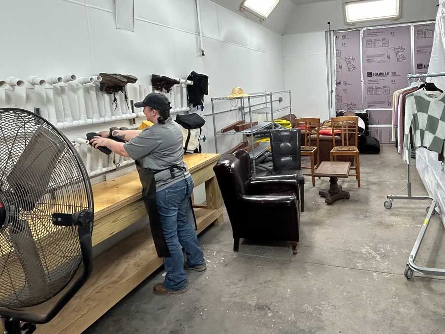 Person working at a workbench in a workshop, with furniture and clothes racks in the background.