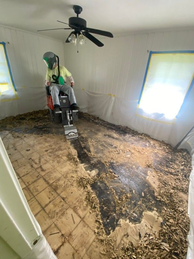 Person using floor scraper in a room, removing old flooring. Protective gear is worn. Walls and windows are covered.