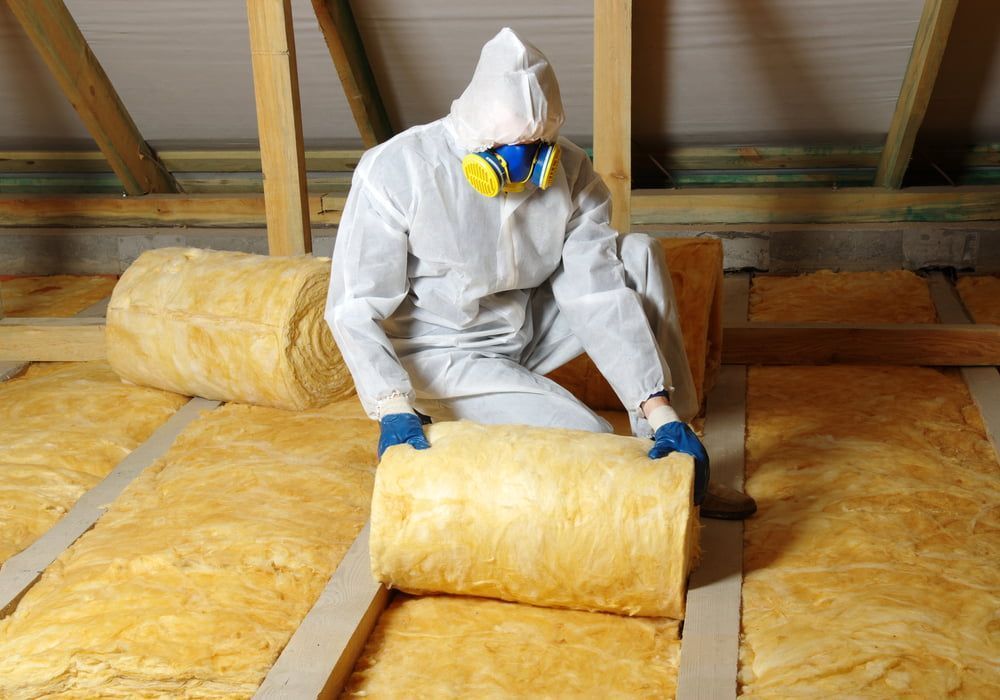 A Man In A Protective Suit Is Sitting On The Floor Holding A Roll Of Insulation — Kosick Roofing NQ Pty Ltd In Mount Low, QLD
