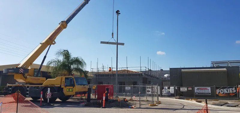 A Yellow Crane Is Lifting A Piece Of Metal In Front Of A Building Under Construction — Kosick Roofing NQ Pty Ltd In Mount Low, QLD