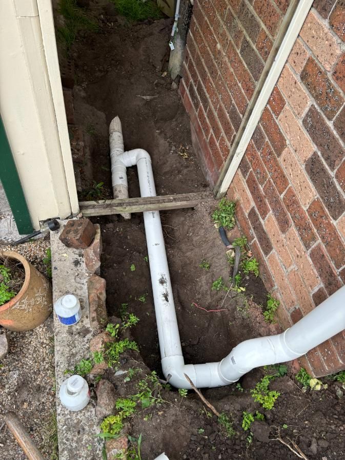 A White Pipe Is Sitting In The Dirt Next To A Brick Wall — Oceanforce Plumbing In Blacksmiths, NSW