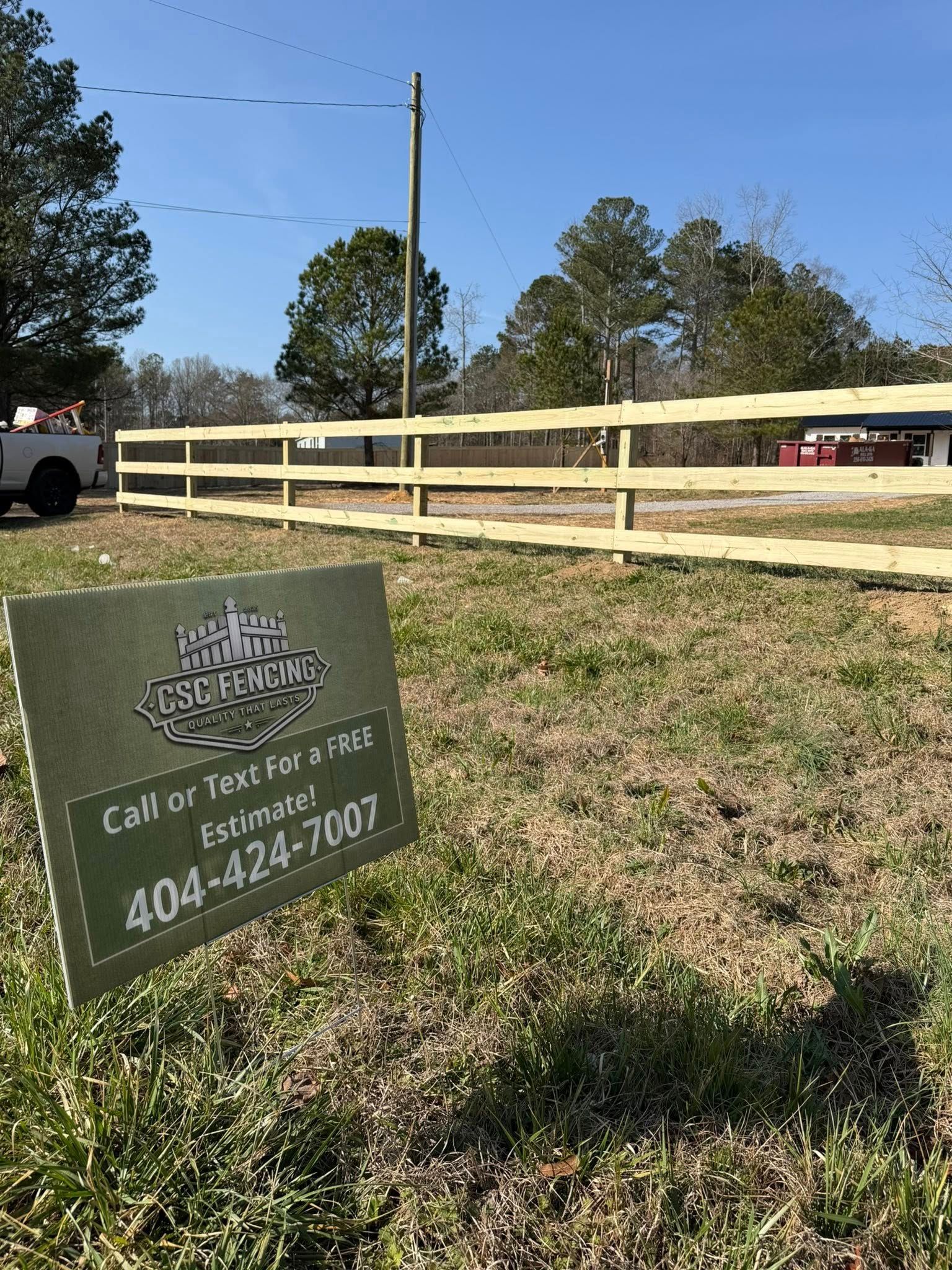 A sign in a grassy field advertising fencing services with the phone number 404-424-7007 in front of a new wooden fence.